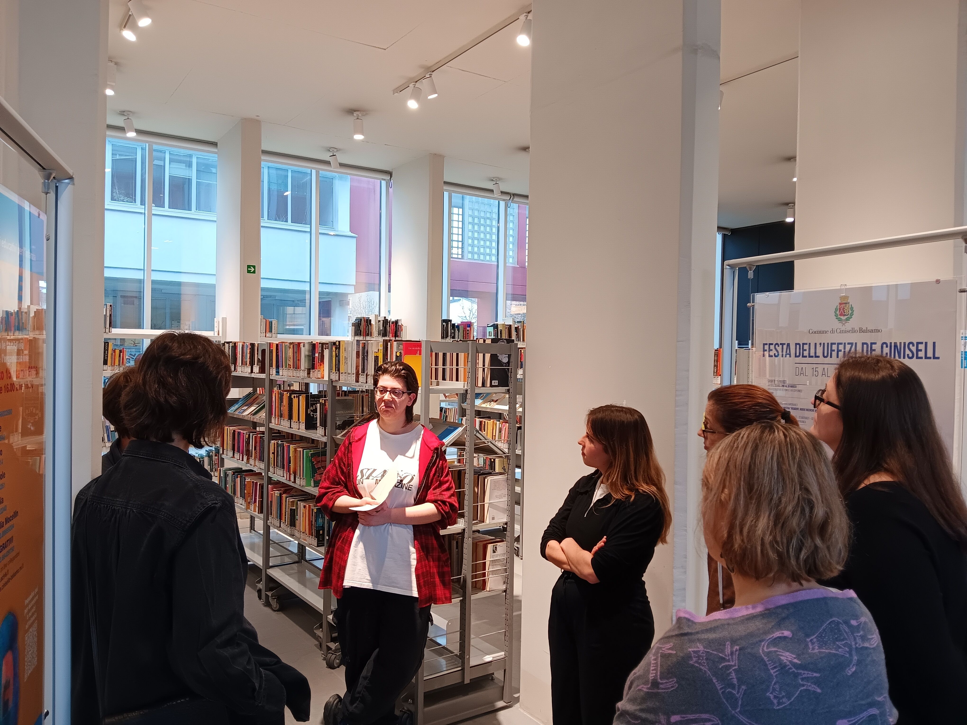 A group of people listens to an informal presentation inside a modern library, surrounded by bookshelves and large windows.