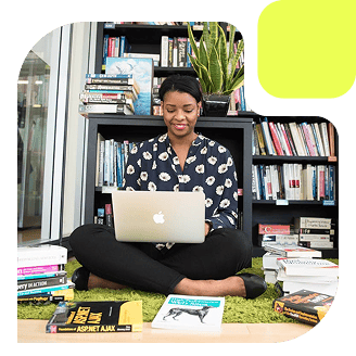Woman sitting on the floor with a laptop, surrounded by books in a bright library.