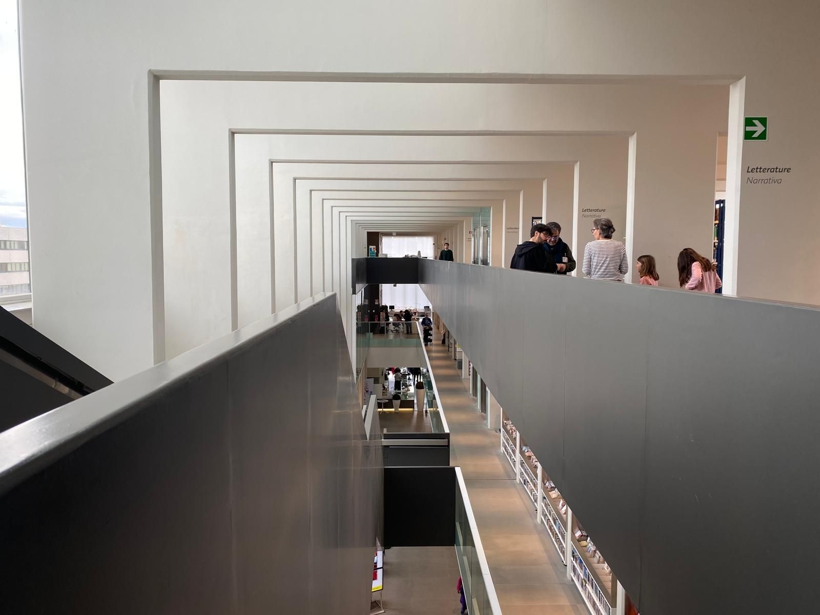 Perspective view of an elevated corridor in a contemporary library, with people exploring the area and bookshelves visible below.