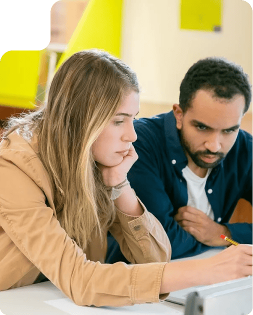 Two young adults focused while working together on a project, seated at a desk with papers and pencils, in a bright and modern environment.