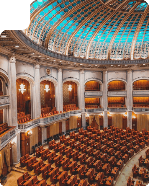 Spacious historic hall with a turquoise glass dome ceiling, wooden furniture arranged in orderly rows, and classical architecture featuring columns, balconies, and elegant chandeliers.